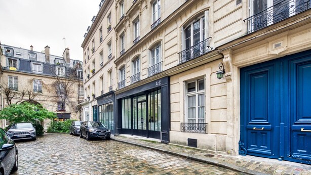 A cobblestone street lined with historic buildings, featuring a black storefront and a blue door. - Blueground | Rambuteau, elevator, nr Chatelet mall (Paris)