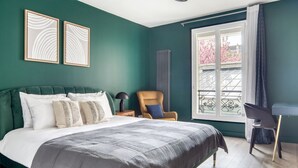 A bedroom with a green wall, a bed with white and gray bedding, two framed artworks, a chair, a desk, and a window with a view of pink trees. - Blueground | Rambuteau, elevator, nr Chatelet mall (Paris)