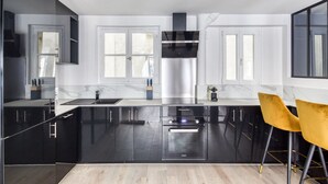 A modern kitchen with black cabinetry, stainless steel appliances, and a yellow chair. - Blueground | Rambuteau, elevator, nr Chatelet mall (Paris)