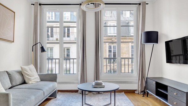A modern living room with a grey sofa, a small round table, a TV on a stand, and large windows with a view of buildings. - Blueground | Louvre, nr Rue Rivoli shops (Paris)