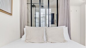 A bedroom with a large bed, two pillows, a blue blanket, and a glass shower door. - Blueground | Louvre, nr Rue Rivoli shops (Paris)