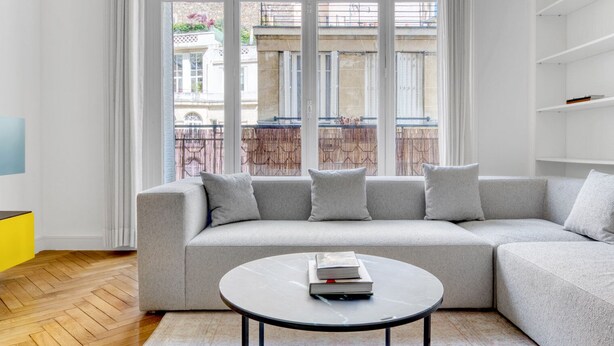 Un salon moderne avec un canapé d’angle gris, une table basse ronde et une vue sur le paysage urbain grâce à de grandes fenêtres.