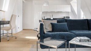 A modern living room with a dark blue sofa, a glass coffee table, and a desk with a chair. A kitchen is visible in the background. - Blueground | Le Marais, renovated, furnished (Paris)