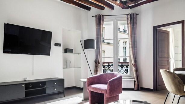 A modern living room with a flat-screen TV, a sofa, a coffee table with books, a floor lamp, and a view of the city through a window with curtains. - Blueground | Le Marais, elev, nr rue de Bretagne (Paris)