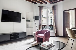 A modern living room with a flat-screen TV, a sofa, a coffee table with books, a floor lamp, and a view of the city through a window with curtains.