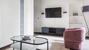 A modern living room with a glass coffee table, a pink armchair, a flat-screen TV mounted on the wall, a dark wooden cabinet, and a floor lamp. - Blueground | Le Marais, elev, nr rue de Bretagne (Paris)