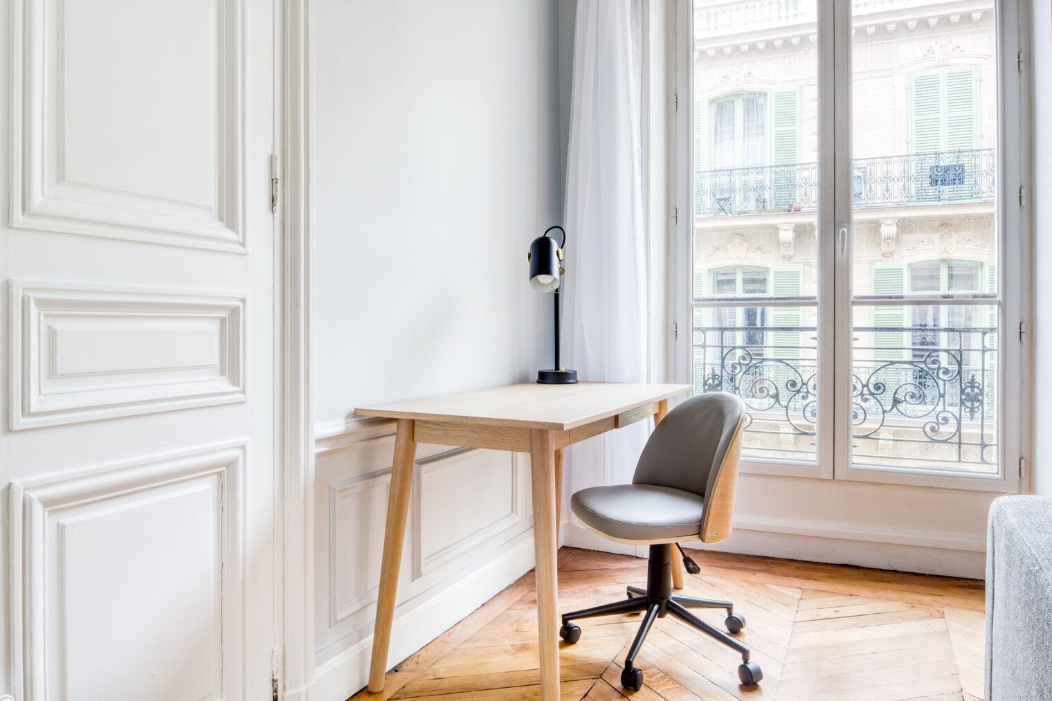 A room with a wooden desk and a grey office chair, a black floor lamp, and a large window with a view of a balcony.