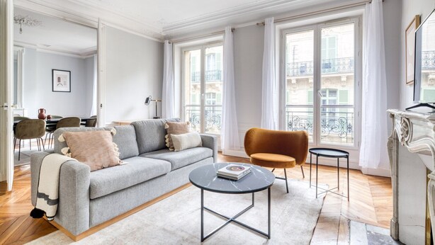 En rummelig stue med en grå sofa, et lille rundt bord og en brun lænestol. Store vinduer med gardiner, et spiseområde i baggrunden, og en balkon er synlig gennem vinduerne.