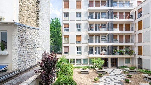 Un immeuble résidentiel à plusieurs étages, doté de balcons, d’une cour avec des sièges et d’un mur en pierre.