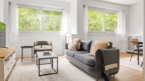 Un salon moderne avec un canapé gris, une petite table, un fauteuil et un téléviseur. Il y a deux fenêtres avec des rideaux, et la vue donne sur un espace verdoyant.