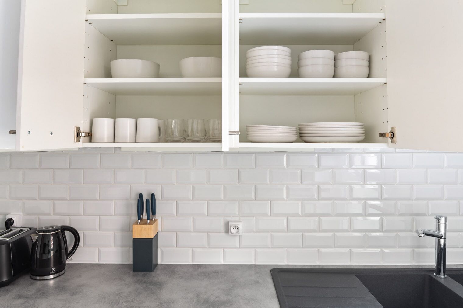 Una cocina con alacenas blancas, salpicadero de azulejos, un lavamanos y una cafetera.