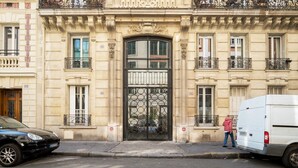 A building with a large ornate entrance, a black gate, and a person standing near a white van. - Blueground | Monceau, furnished, near park (Paris)