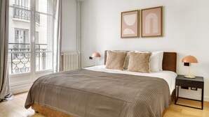 Een slaapkamer met een groot bed, twee bedlampen, een radiator en een raam met uitzicht op een balkon.