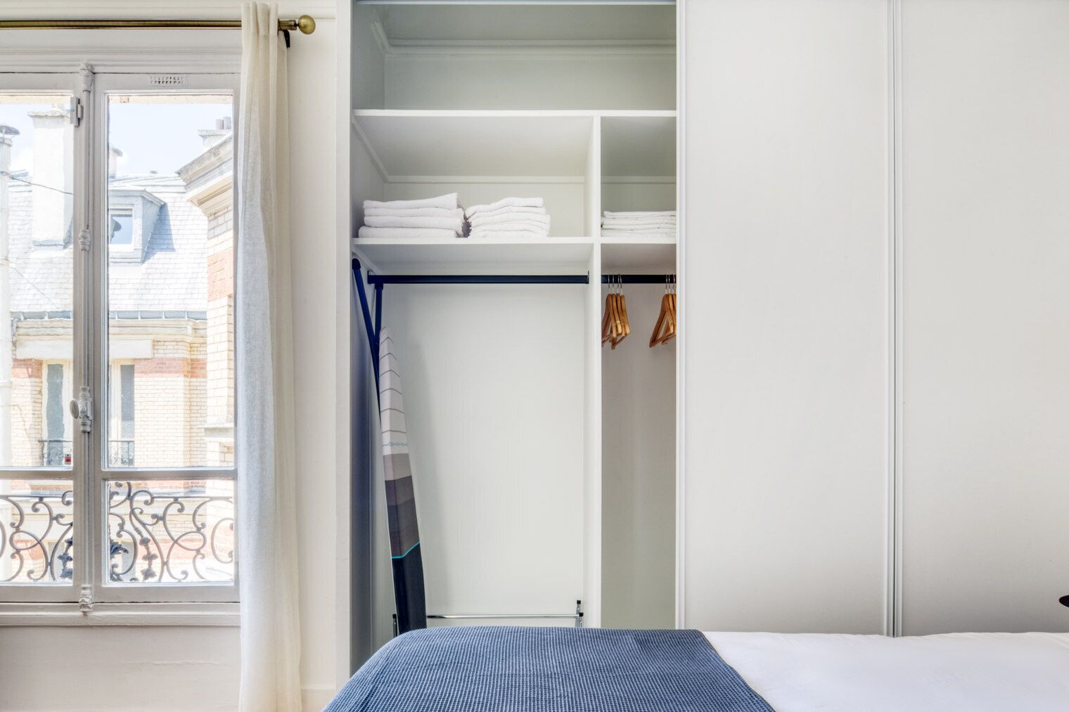 Ein Schlafzimmer mit einem Bett, einem weißen Kleiderschrank mit Einlegeböden und einem Fenster mit Blick auf Gebäude.