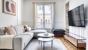 Een moderne woonkamer met een grijze bank, een ronde salontafel, een aan de muur bevestigde televisie en uitzicht op de straat door een groot raam.