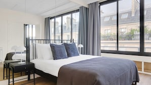 A modern hotel room with a large bed, a bedside table, a lamp, and a view of the city through large windows. - Blueground | Trocadéro, nr Eiffel Tower (Paris)