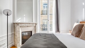 Une chambre à coucher avec un lit, une cheminée, un balcon avec une balustrade et une vue sur les bâtiments environnants.
