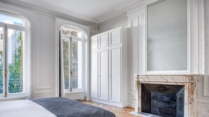 Une chambre avec un grand lit, une cheminée et une vue sur la verdure par la fenêtre.