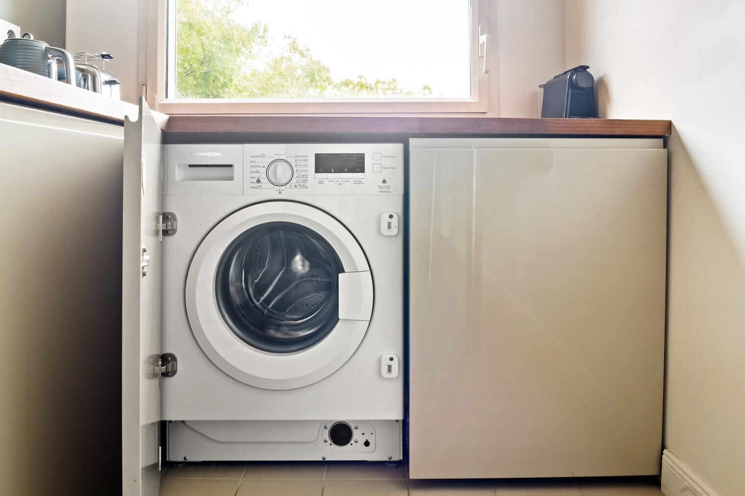 A compact, modern washing machine in a small laundry room with a window above it.