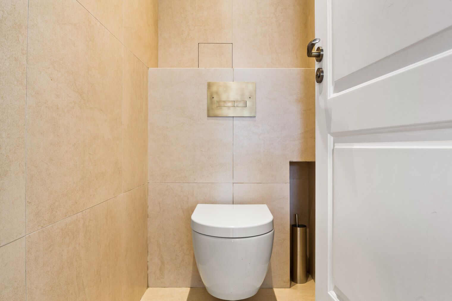 A modern bathroom with a white toilet, a wall-mounted flush handle, and a door with a metallic handle.