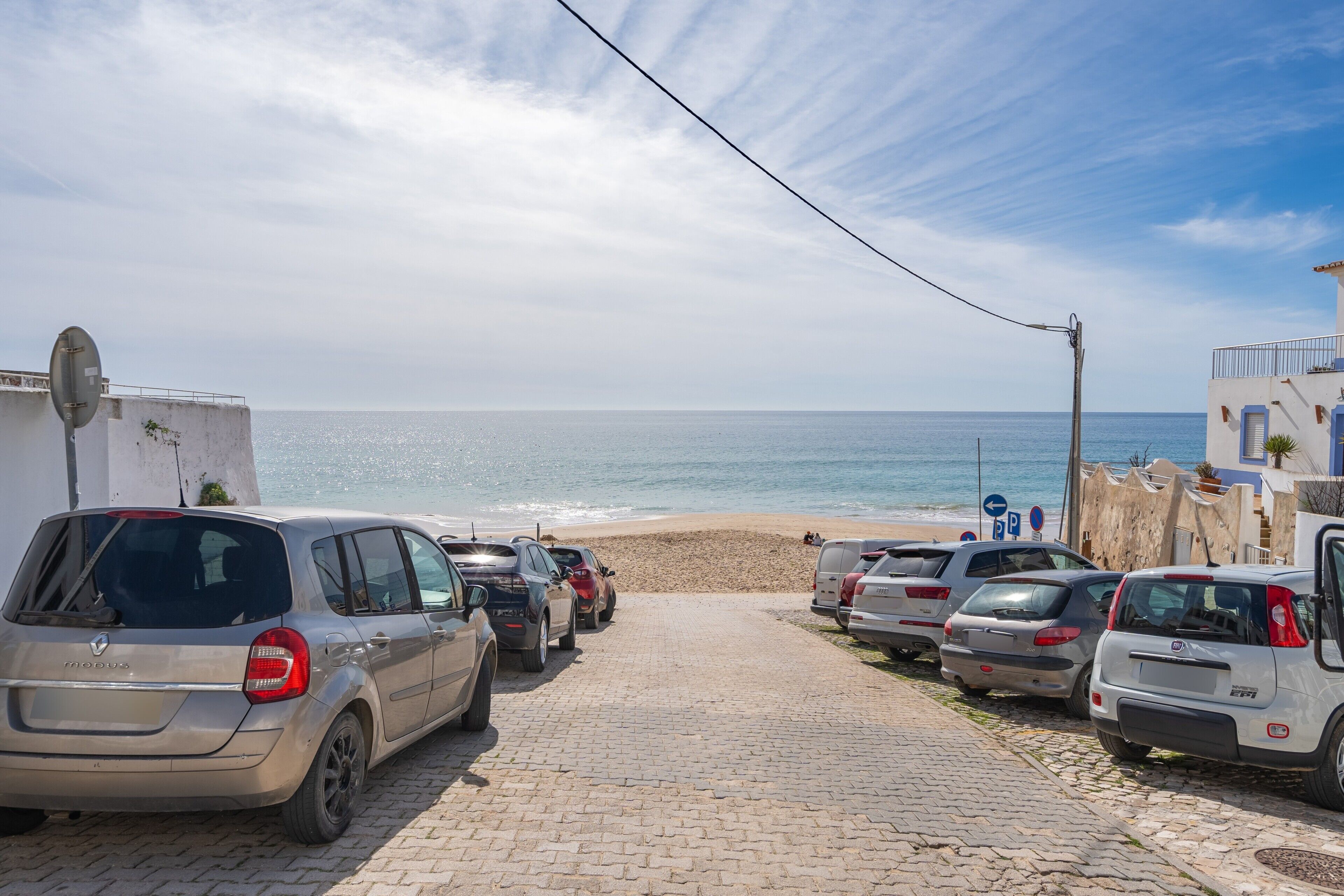 En asfalteret sti der fører til en strand, med parkerede biler og frit udsyn til havet.