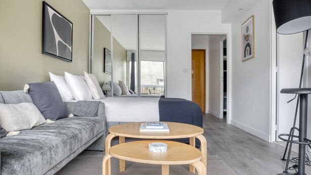 A modern hotel room with a grey sofa, a round wooden table, a large bed, a mirror, and a black floor lamp. - Blueground | Auteuil, balcony, nr Parc de Princes (Paris)