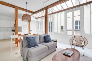 A modern living room with a grey sofa, wooden beams, large windows, and a dining area with a white table and chairs. - Blueground | Raspail - Montparnasse, furnished (Paris)