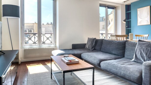 A modern living room with a grey sofa, a wooden coffee table, and a large window with a view of buildings. - Blueground | Sentier, furnished, nr Montorgueil St (Paris)