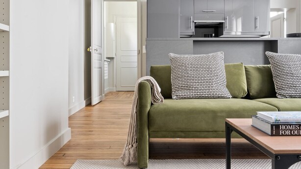 Un salon moderne avec un canapé vert, des coussins à motifs et une table basse sur laquelle repose un livre. On aperçoit à l’arrière-plan une cuisine avec des meubles gris et une porte blanche.