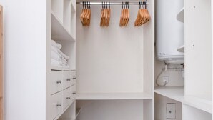 Un armario vestidor blanco con estantes abiertos, una barra para colgar con perchas de madera y un lavamanos blanco con espejo.