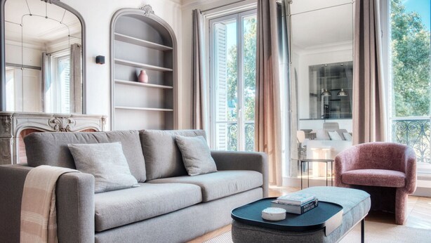 Una sala de estar moderna con un sofá gris, una mesita redonda y un sillón rosa. Hay una estantería y un ventanal con cortinas.