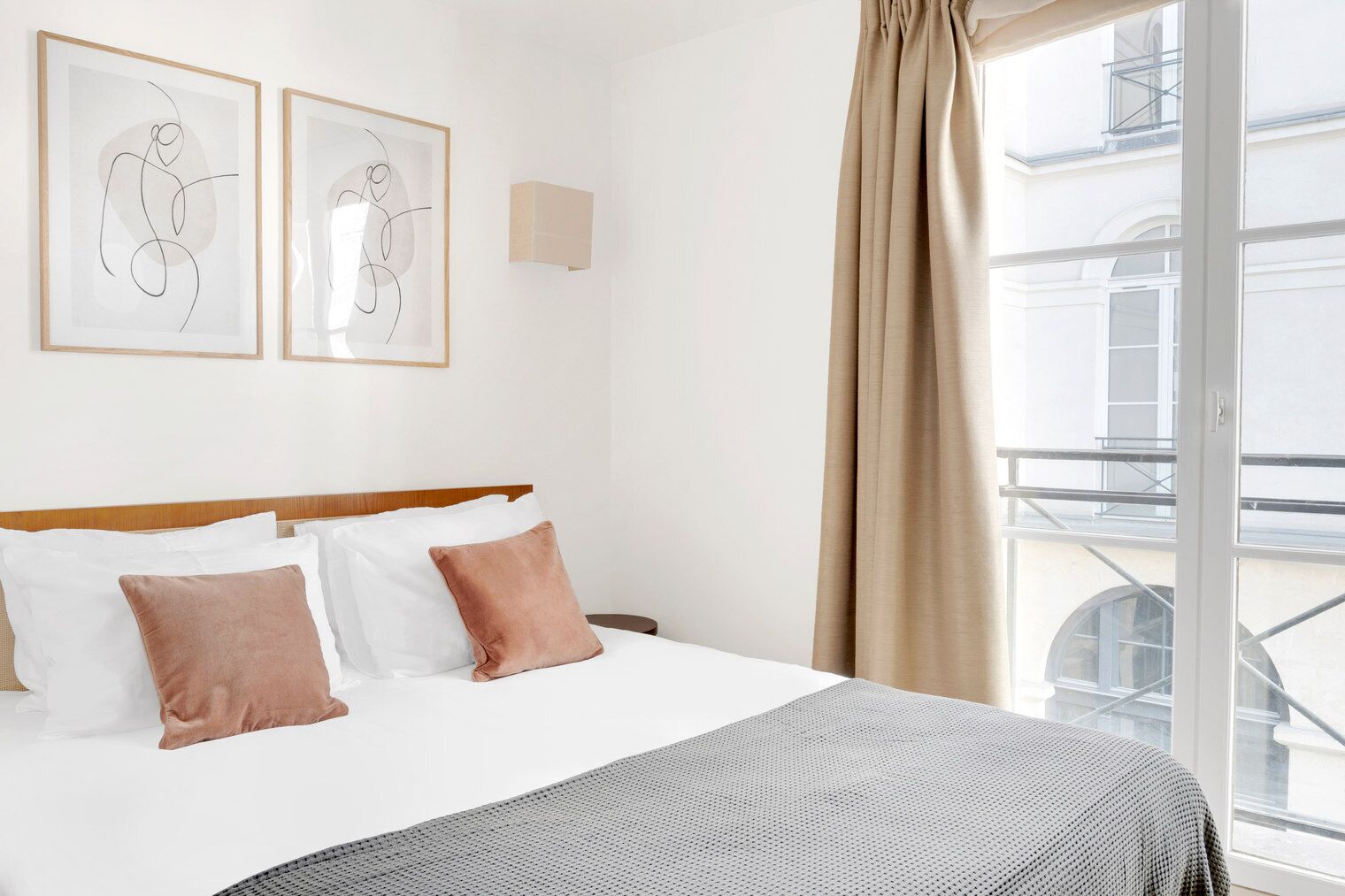 Habitación de hotel con cama, dos obras de arte abstractas enmarcadas, una ventana con vista a un edificio y un balcón.