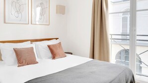 A hotel room with a bed, two framed abstract artworks, a window with a view of a building, and a balcony.