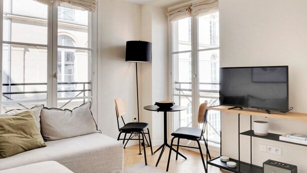 A modern, well-lit room with a sofa, two chairs, a small table, a TV, and a balcony with large windows.
