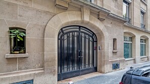 Une façade d’immeuble avec une grande entrée en arc de cercle, une petite fenêtre ornée d’une plante et une voiture garée dans la rue.