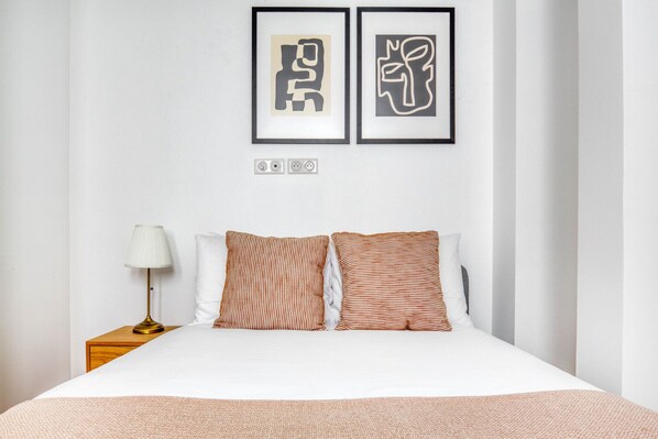 Ein ordentlich bezogenes Bett mit gestreiften Kissen, eine Nachttischlampe und zwei abstrakte Kunstwerke an der Wand.