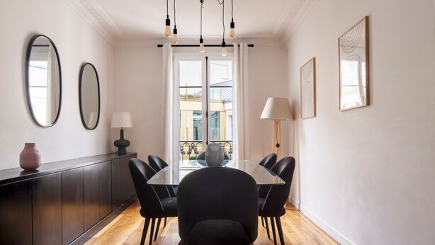 Un coin repas comprenant une table ronde en verre, six chaises noires, un buffet en bois foncé, deux miroirs muraux, un lampadaire et une vue sur le balcon extérieur.