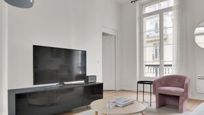 A modern living room with a large flat-screen TV, a black entertainment unit, a round wooden coffee table, a pink armchair, and a view of the city through large windows. - Blueground | Sentier, balcony, nr Les Halles (Paris)