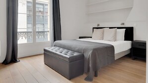A modern bedroom with a large bed, a grey upholstered bench, a nightstand, and a window with curtains. - Blueground | Sentier, balcony, nr Les Halles (Paris)