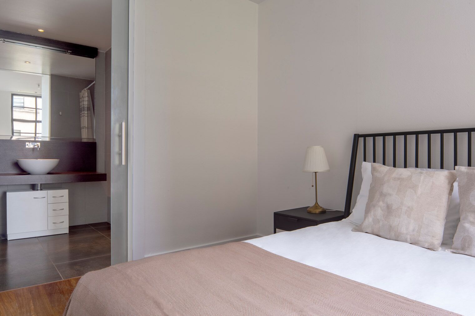 Un dormitorio moderno con una cama grande, una mesita de noche con lámpara, y un baño visible a través de una puerta abierta.