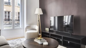 A modern living room with a large flat-screen TV, a white armchair, a floor lamp, and a round coffee table with books and a decorative item.