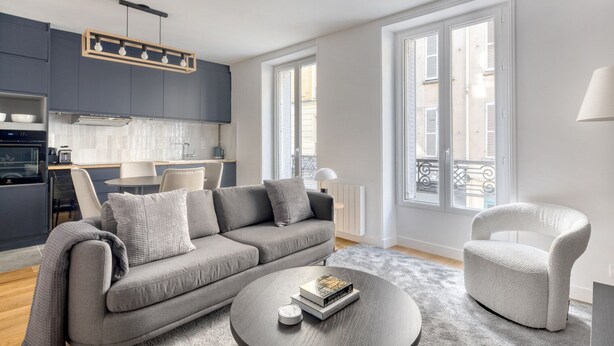 A modern living room with a grey sofa, a round coffee table, and a white armchair. There is a kitchen area with grey cabinets and a dining table. Large windows provide natural light and a view of the outside.