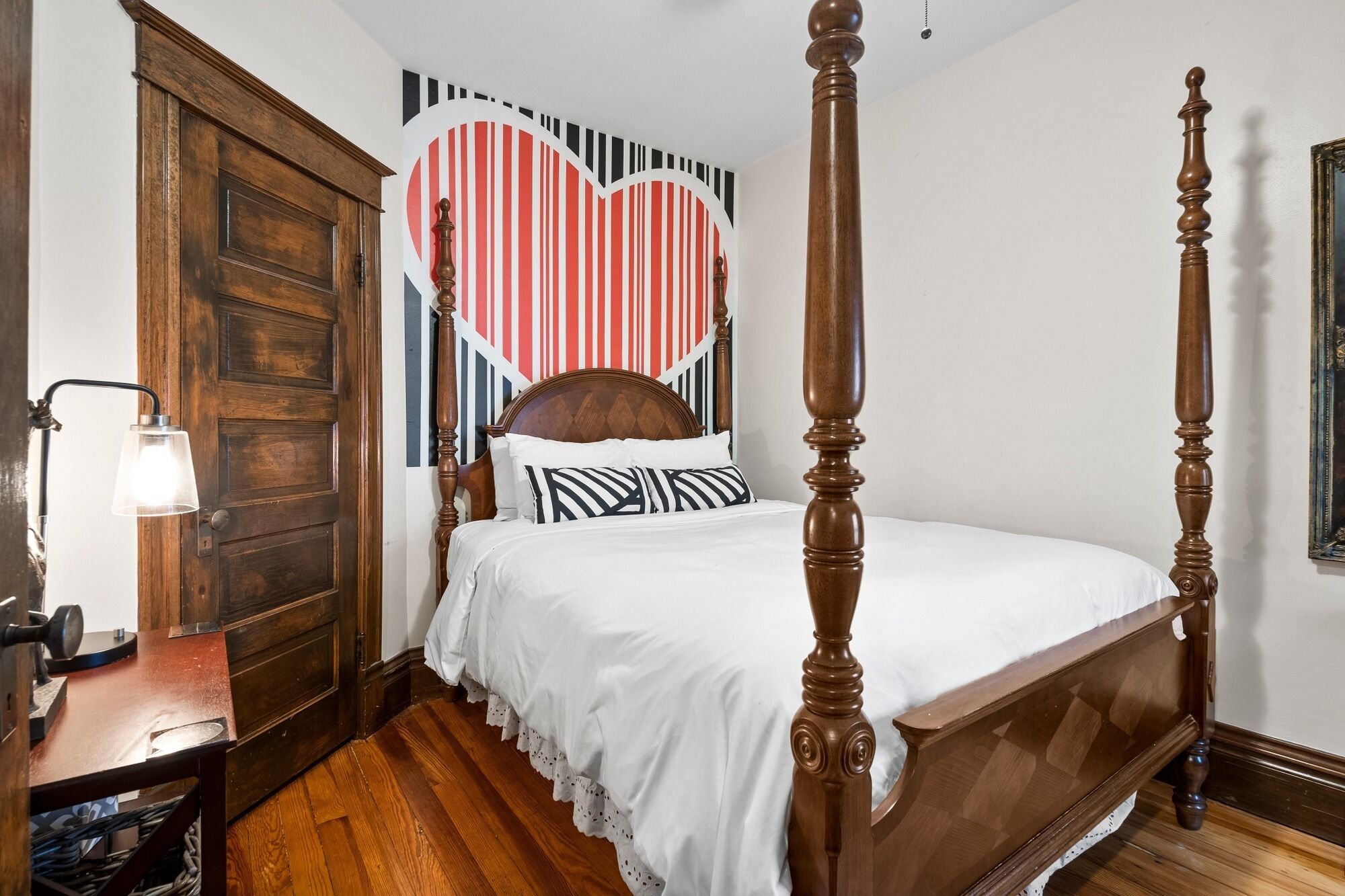 A four-poster bed with a heart-shaped wall decoration, a wooden nightstand with a lamp, and a wooden floor.