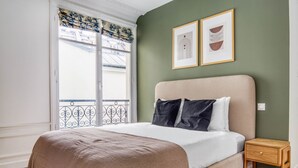 A bedroom with a bed, a nightstand, a window with curtains, and two framed pictures on the wall. - Blueground | Le Marais, elevator, nr Rue Rambuteau (Paris)