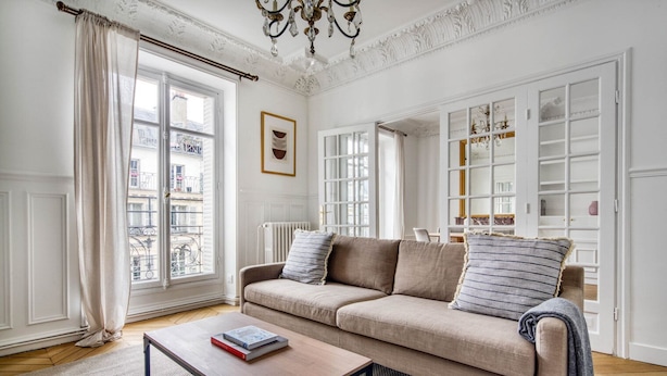 A spacious living room with a beige sofa, a wooden coffee table, and a view of the city through large windows. - Blueground | Le Marais, elevator, nr Rue Rambuteau (Paris)