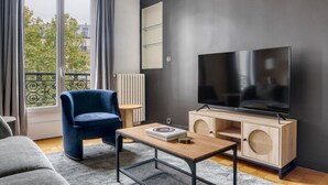 A modern living room with a large flat-screen TV, a blue armchair, a wooden coffee table, and a view of the city through large windows.