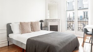 A bedroom with a bed, a fireplace, a balcony with a railing, and a view of buildings. - Blueground | Bir Hakeim, nr Eiffel Tower (Paris)