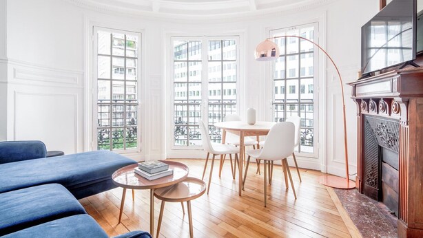 A spacious living room with a blue sofa, a round table with books, and a fireplace. Large windows offer views of buildings outside. - Blueground | Bir Hakeim, nr Eiffel Tower (Paris)