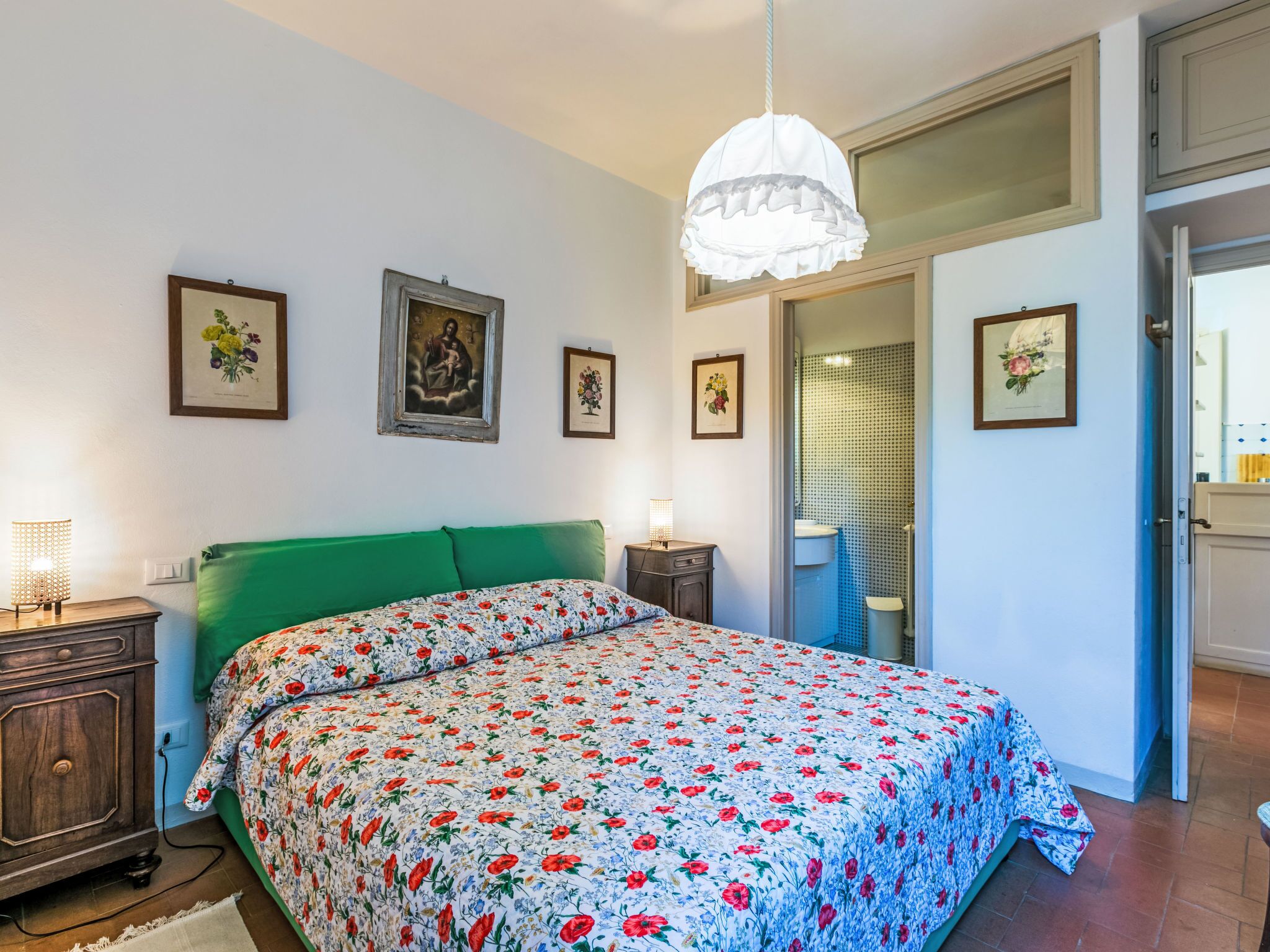 A bedroom with a floral bedspread, a green headboard, a bedside table with a lamp, and framed artwork on the wall.