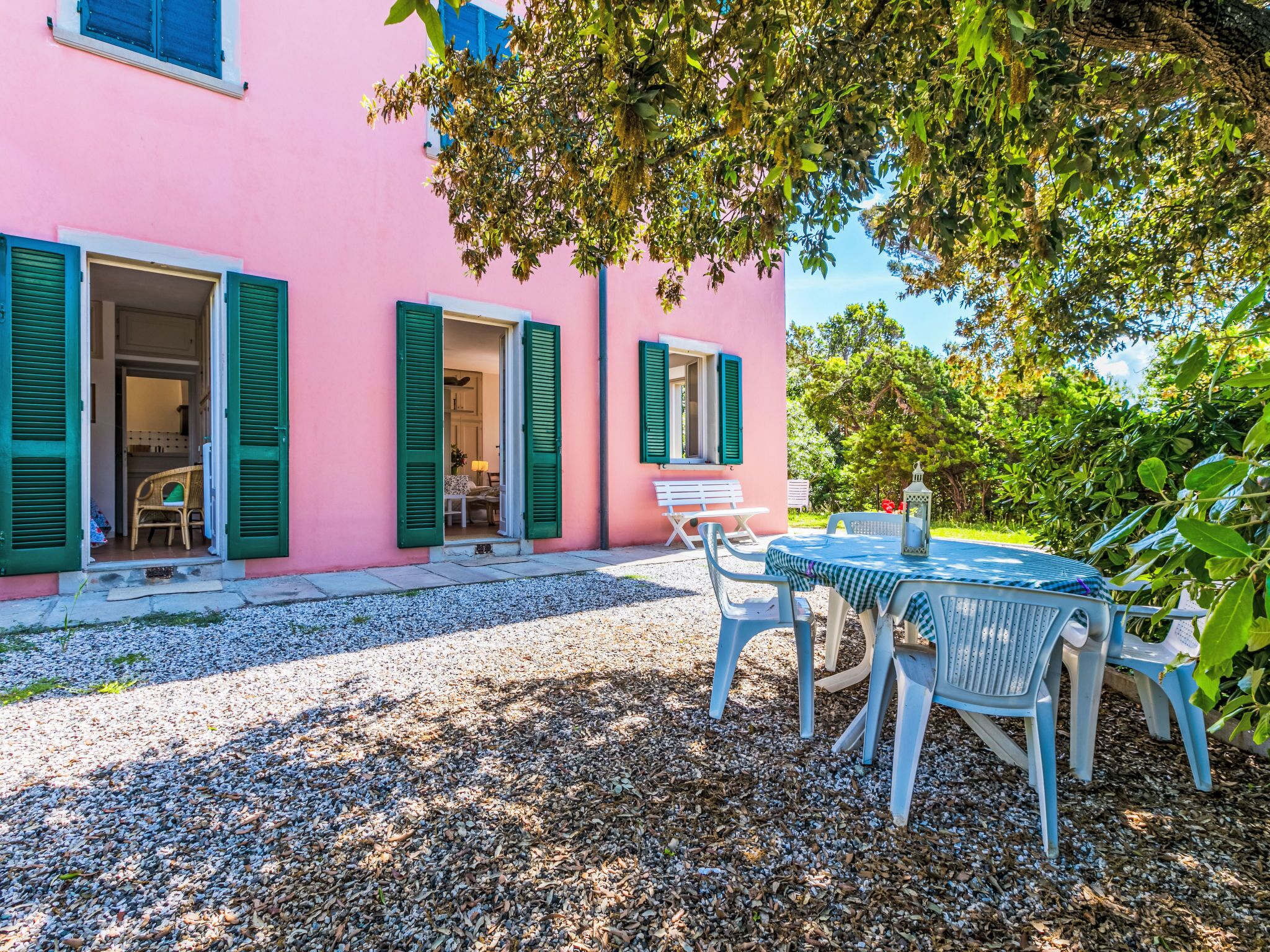A pink building with green shutters, a gravel path, and a table with chairs outside.